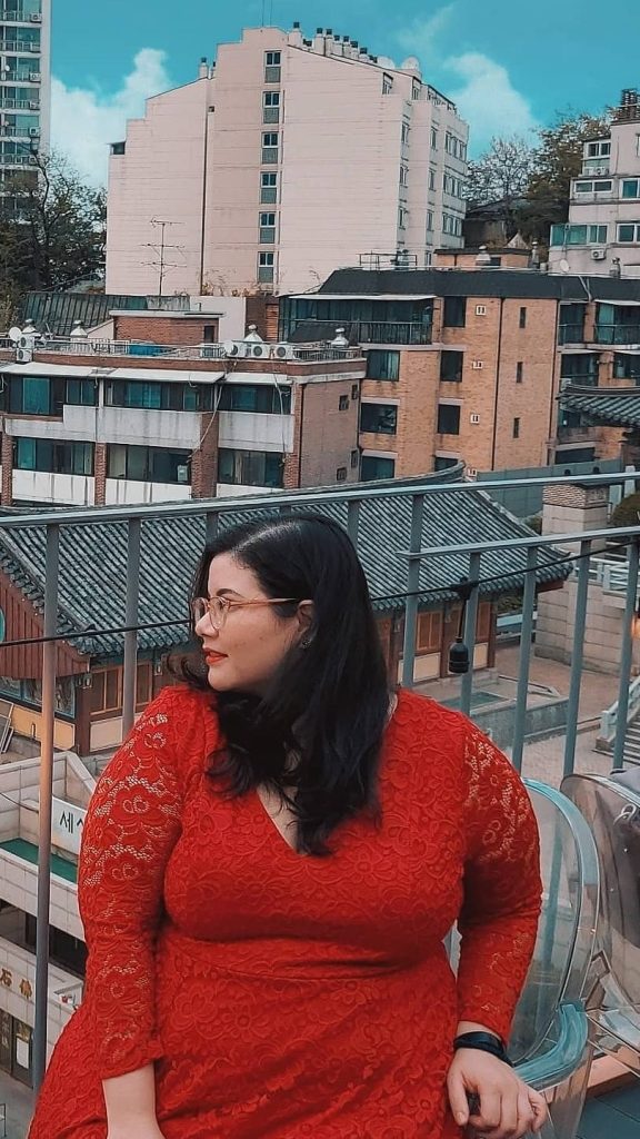 Plus size woman with black hair and glasses sits on a chair in a red dress. She is seated outside on a balcony of a popular book café. The backdrop is the Buddhist temple adjacent to the book café. There are also apartment buildings and other housing in the background. This café can be found in Mapo-gu, Seoul.